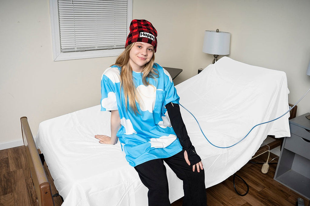 Young girl wearing a blue cloud-patterned adaptive shirt and a red plaid beanie, sitting on a bed.