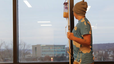 Person wearing a beanie and hospital gown holding an IV stand, looking out a window at a cityscape.
